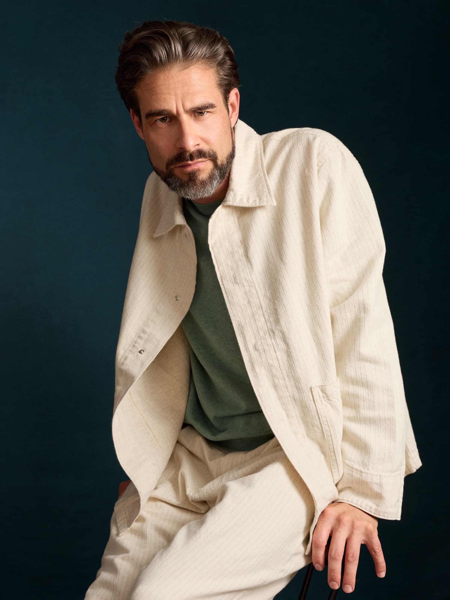 Brandon Mills — three-quarter seated portrait in a cream overshirt over sage tee against a teal backdrop