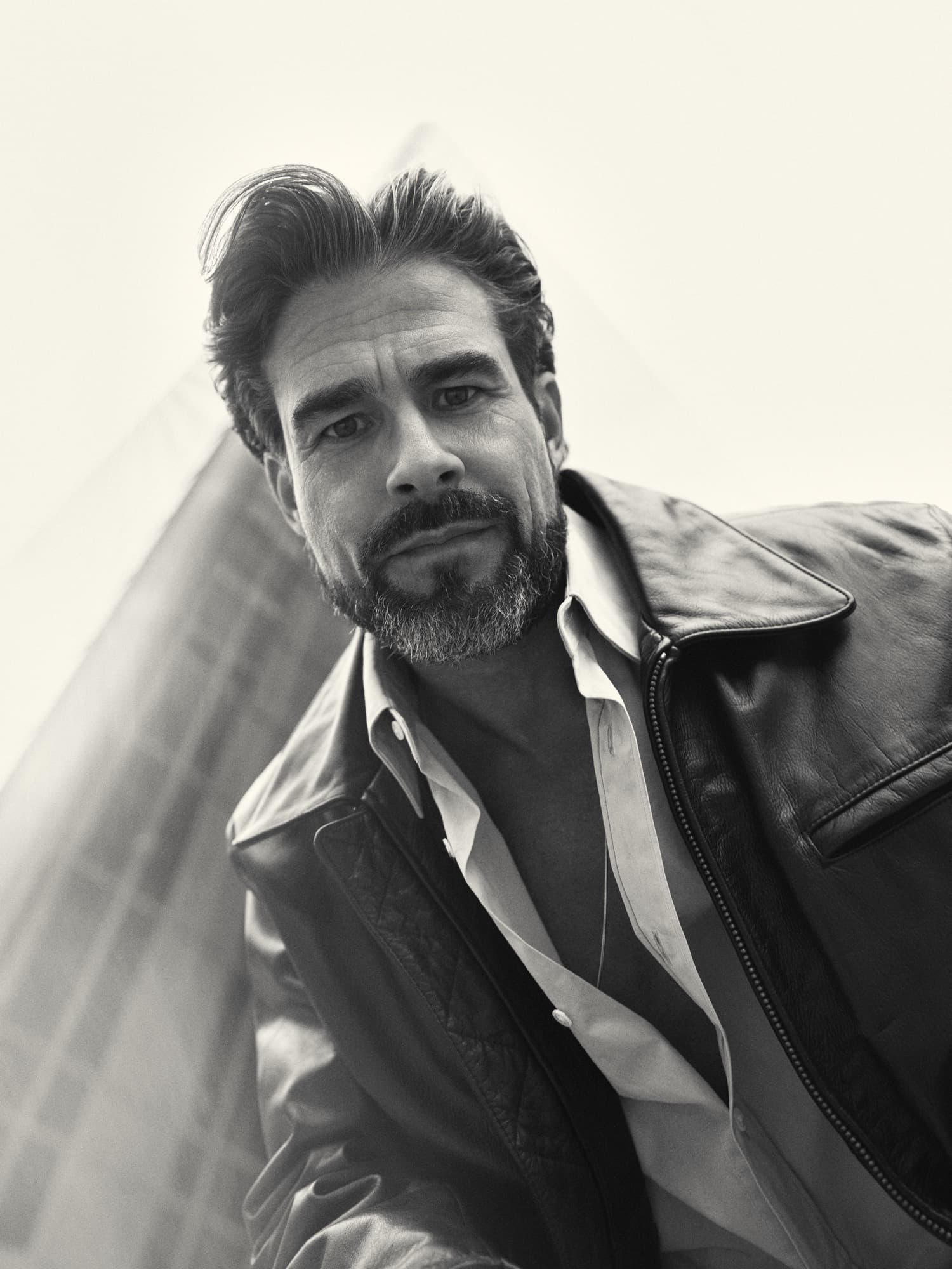 Brandon Mills — low-angle black-and-white editorial portrait in a leather jacket against an architectural backdrop